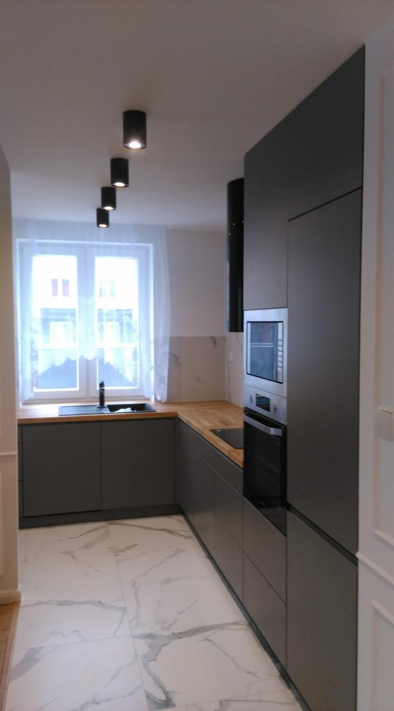 Nowoczesna kuchnia w odcieniach szarości z drewnianym blatem, białymi marmurowymi płytkami podłogowymi i rzędem czarnych lamp sufitowych.