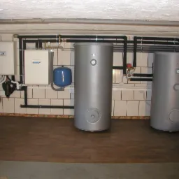 Dwa srebrne zasobniki ciepłej wody w kotłowni z brązową podłogą i białymi płytkami ściennymi, rury w izolacji termicznej, manometry na zbiornikach, widok z niskiego kąta.