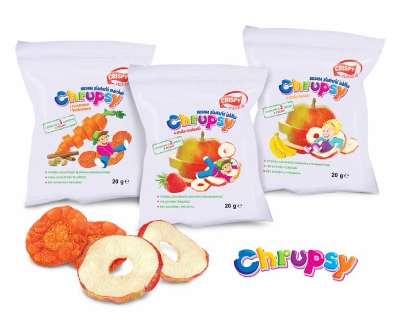 Trzy białe opakowania suszonych plasterków owocowych i warzywnych marki Chrupsy: marchew z kardamonem, jabłko z truskawką i jabłko z bananem. Na pierwszym planie suszone plasterki marchewki i jabłka.
