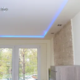 Biały sufit napinany z niebieskim oświetleniem LED obwodowym i dwoma punktowymi lampami, ściana wykończona dekoracyjną okładziną imitującą kamień, widok na drzewo za oknem.