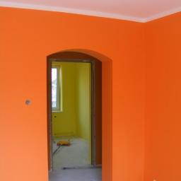 Lar-Bed - Pomarańczowe ściany w pokoju po malowaniu, widoczny fragment sufitu z listwą, przejście do pokoju pomalowanego na żółto, wałek do malowania na podłodze, puszka po farbie w rogu.