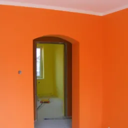 Pomarańczowe ściany w pokoju po malowaniu, widoczny fragment sufitu z listwą, przejście do pokoju pomalowanego na żółto, wałek do malowania na podłodze, puszka po farbie w rogu.