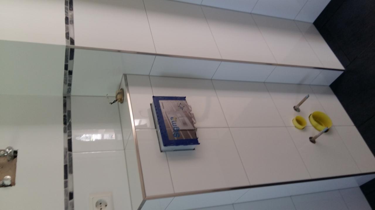 Białe płytki ścienne z metalowymi listwami wykończeniowymi, widoczne otwory na instalacje, puszka elektryczna, karton po narzędziach i żółte wiaderka na podłodze, w trakcie prac wykończeniowych...