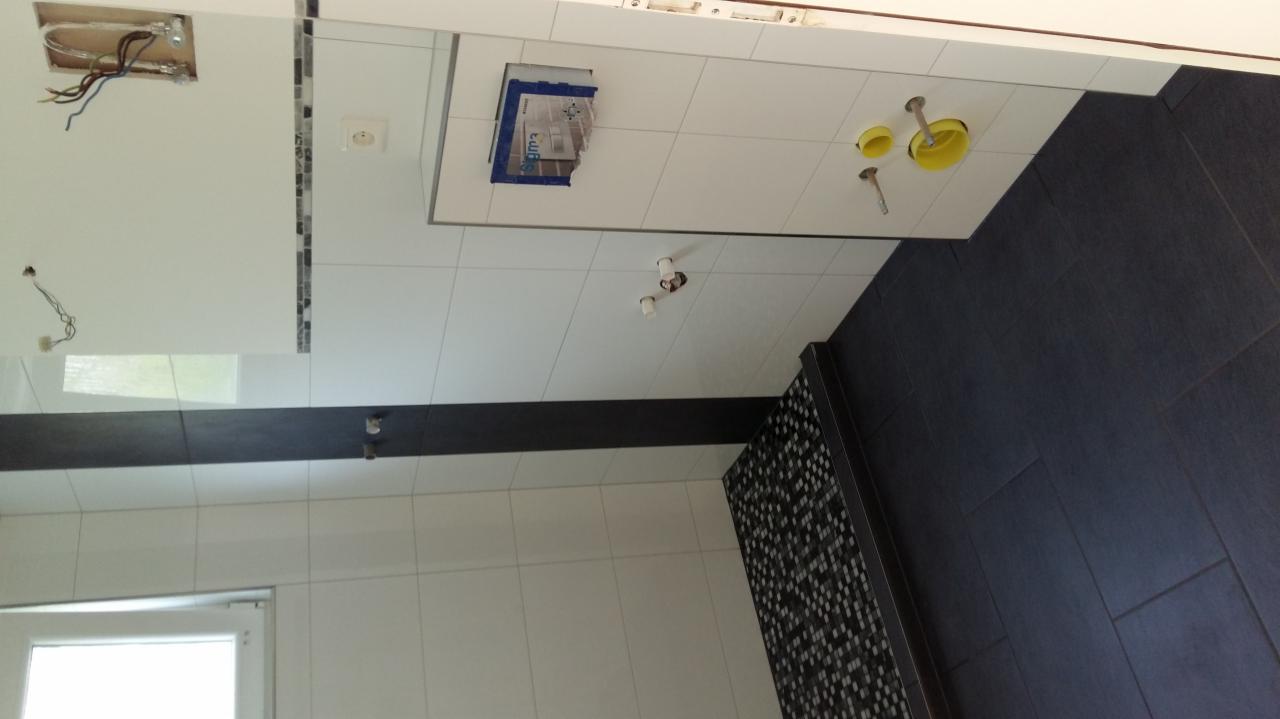 Wnętrze łazienki w trakcie remontu, widoczne białe płytki ścienne, fragment czarnej podłogi z płytek, instalacja sanitarna w ścianie oraz wystające przewody elektryczne.