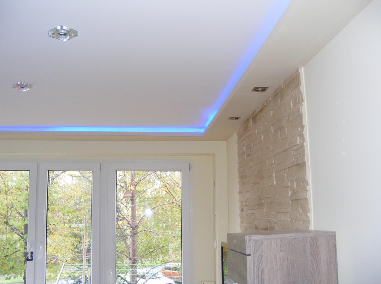 Biały sufit napinany z niebieskim oświetleniem LED obwodowym i dwoma punktowymi lampami, ściana wykończona dekoracyjną okładziną imitującą kamień, widok na drzewo za oknem.