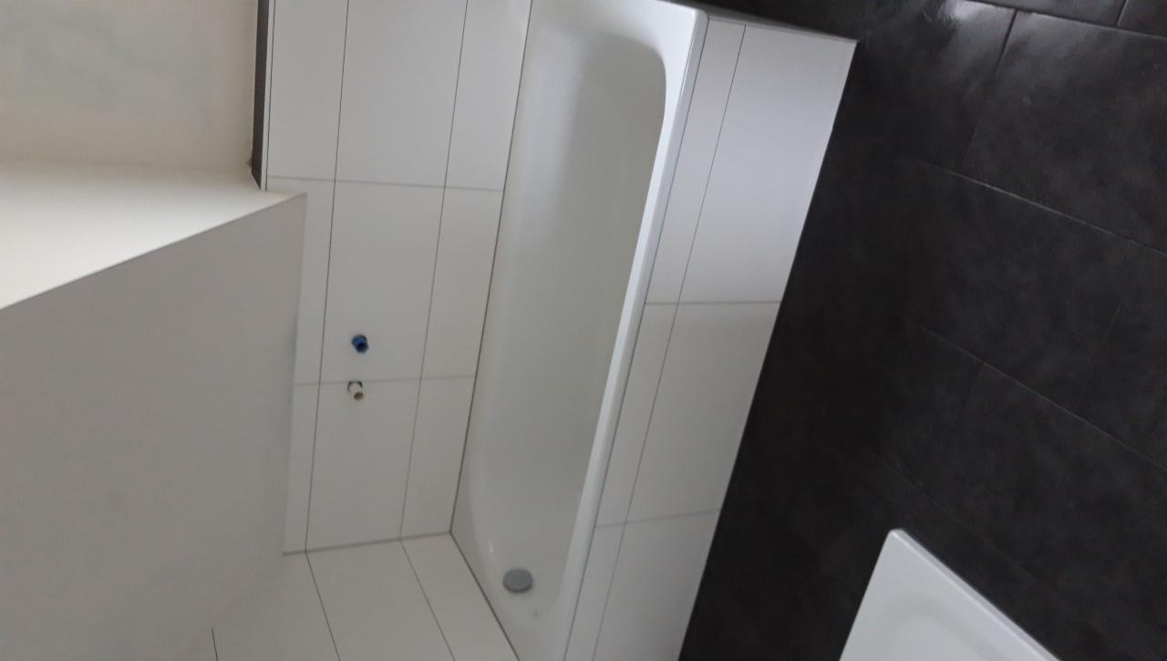 Wanna obudowana białymi płytkami, na ścianie widoczne przyłącza wodne, obok ciemne płytki imitujące kamień.