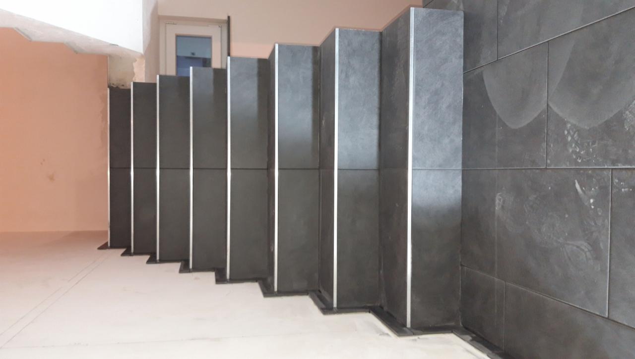 Wyłożone ciemnymi płytkami schody w trakcie prac wykończeniowych, widoczne niedociągnięcia i ślady po zaprawie na płytkach oraz niezabezpieczone krawędzie schodów.