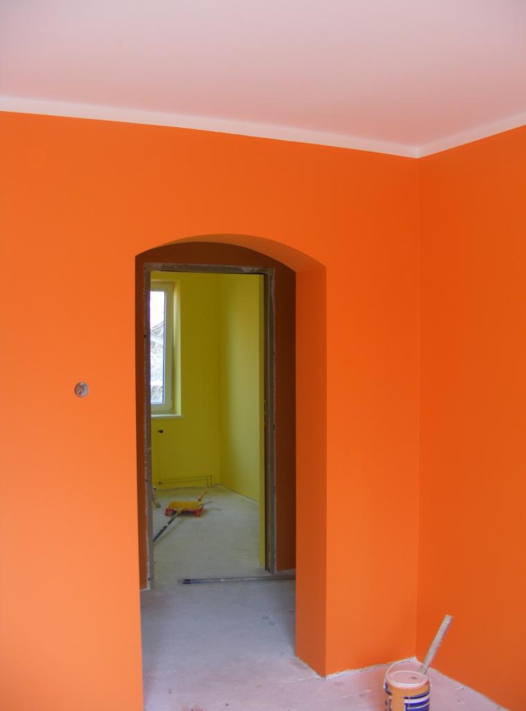 Pomarańczowe ściany w pokoju po malowaniu, widoczny fragment sufitu z listwą, przejście do pokoju pomalowanego na żółto, wałek do malowania na podłodze, puszka po farbie w rogu.