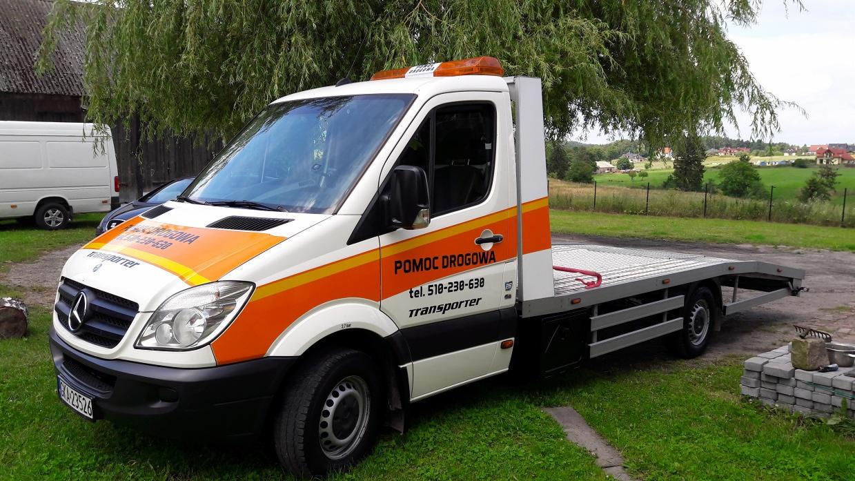 Biało-pomarańczowy Mercedes Sprinter z napisem 'Pomoc Drogowa' zaparkowany na trawie, widok z boku, z tyłu sterta szarych kostek brukowych i narzędzia.