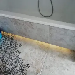 Fragment wykończonej łazienki z wanną, dekoracyjnymi płytkami podłogowymi w czarno-białe wzory oraz podświetleniem LED pod wanną.