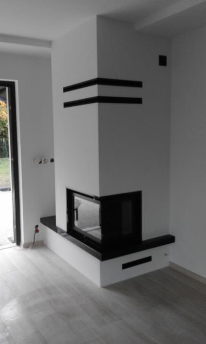Biały narożny wkład kominkowy z czarnymi akcentami w nowoczesnym wnętrzu, widoczny z boku. Minimalistyczny design z prostymi liniami i gładkimi powierzchniami.