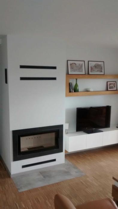 Nowoczesny wkład kominkowy z czarną ramą, wbudowany w białą ścianę z poziomymi szczelinami wentylacyjnymi, na tle drewnianej podłogi i białych mebli z półkami.