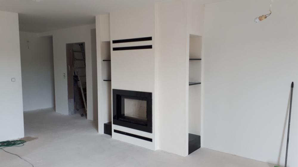 Nowoczesny, biały kominek z czarnymi akcentami w surowym wnętrzu, widoczne niedokończone wykończenia ścian i podłogi, drabina w tle sugeruje trwające prace.