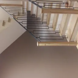 Nowoczesne, drewniane schody z czarną, metalową balustradą, widok od dołu, z oświetleniem sufitowym i fragmentem drewnianej podłogi.