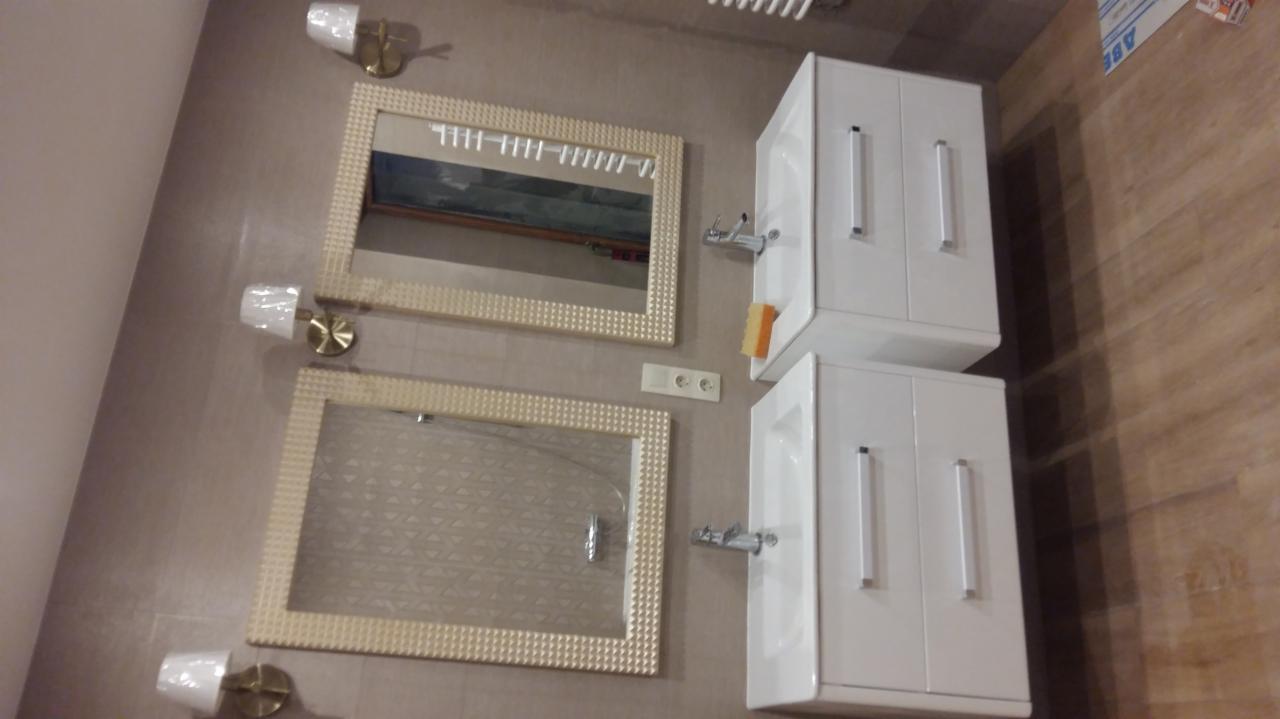 Dwie białe szafki pod umywalki z chromowanymi uchwytami, zawieszone obok dwóch kwadratowych luster w ozdobnych, beżowych ramach, oświetlone przez cztery kinkiety z białymi abażurami i złotymi...