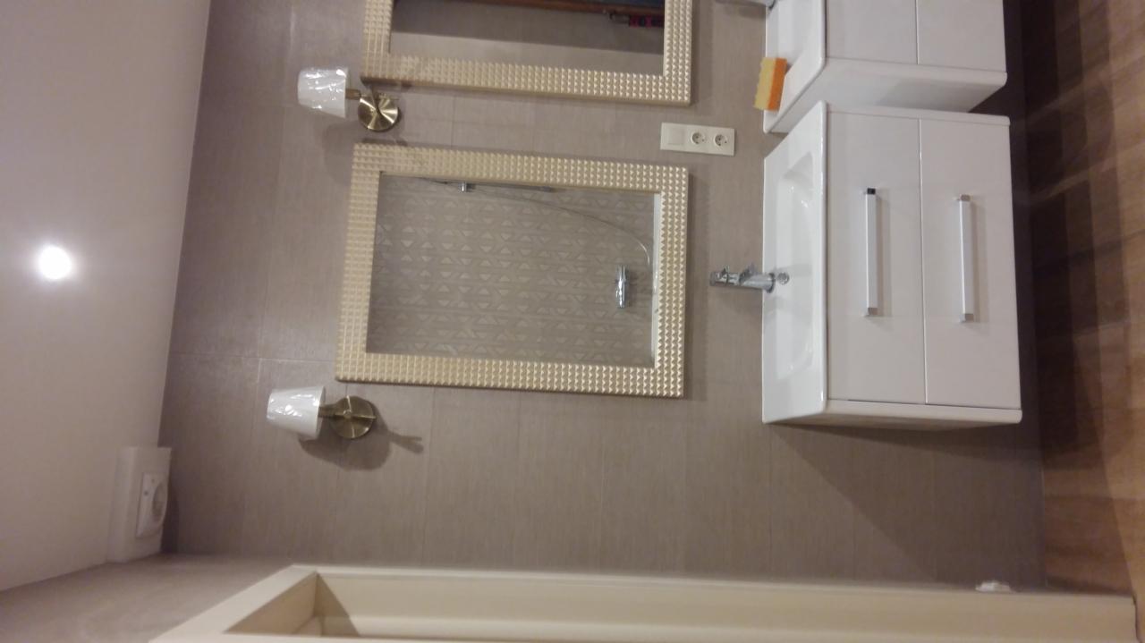 Nowoczesna łazienka z dwoma lustrami w ozdobnych ramach, białą szafką pod umywalką i brązowymi płytkami na ścianie, oświetlona lampami ściennymi.