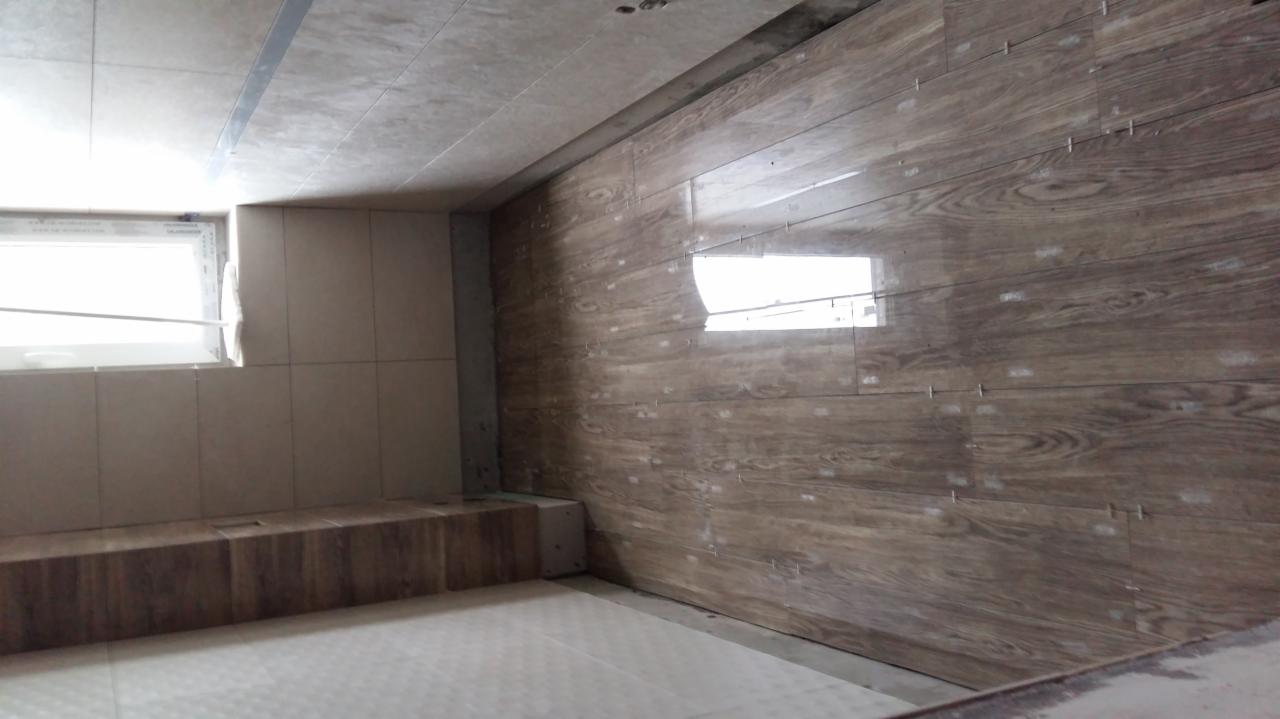 Ściana w trakcie układania płytek imitujących drewno w łazience, widoczne krzyżyki dystansowe, jasne płytki podłogowe i okno.