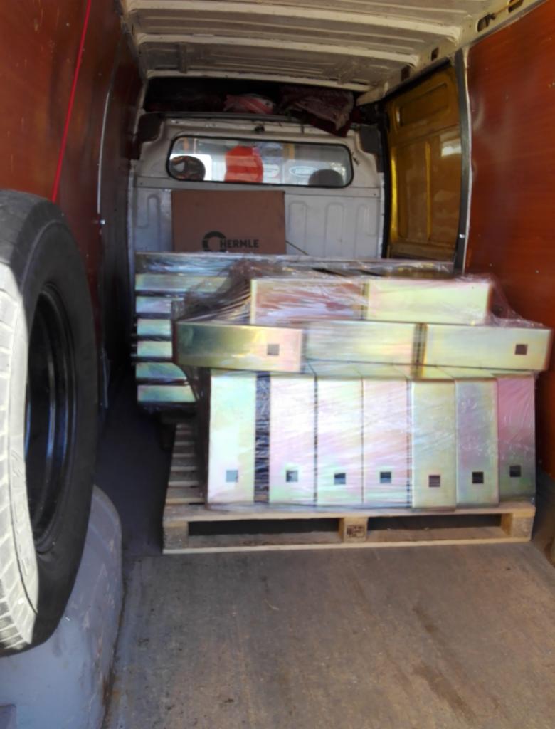 Wnętrze furgonetki z załadowanym paletą metalowych elementów zabezpieczonych folią stretch, widoczna opona i karton z logo Hermle.