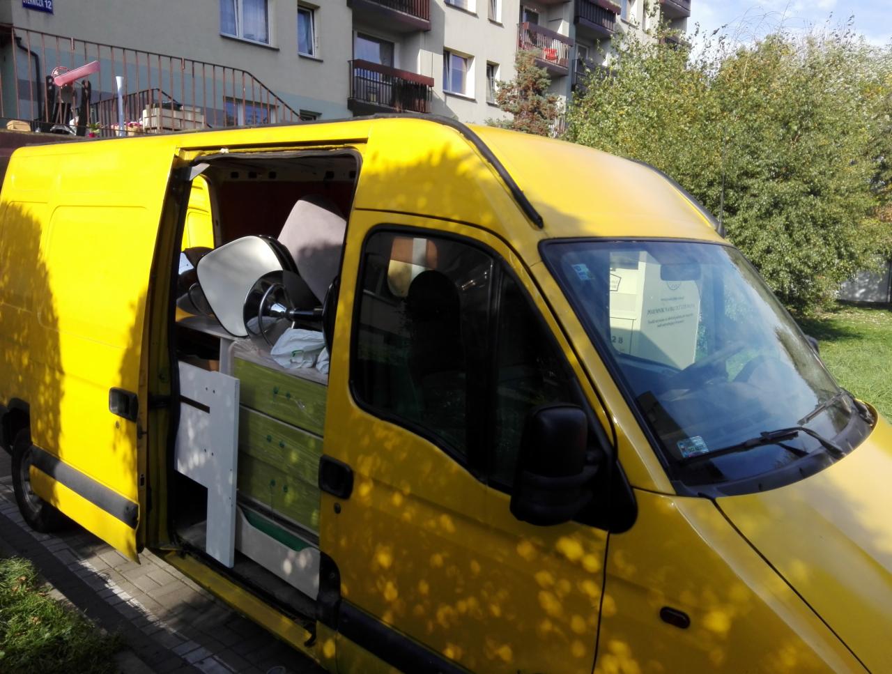 Żółty bus z otwartymi drzwiami bocznymi, załadowany meblami biurowymi i elementami wyposażenia wnętrz, zaparkowany na chodniku przed budynkiem mieszkalnym.