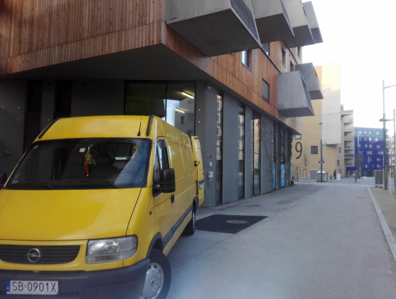 Żółty Opel Movano zaparkowany przed nowoczesnym budynkiem z drewnianą elewacją i betonowymi balkonami. Na szybie widać zawieszkę. Tablica rejestracyjna SB 0901X.