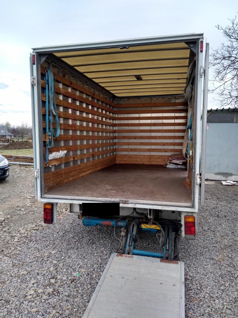 Wnętrze paki transportowego busa z drewnianą zabudową ścian, widok z tyłu, rampa załadunkowa opuszczona, niebieskie pasy mocujące po lewej stronie.