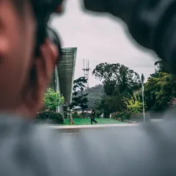 Osoba fotografująca osobę biegnącą chodnikiem w parku miejskim, z wieżą radiową w tle, widziana przez obiektyw aparatu.