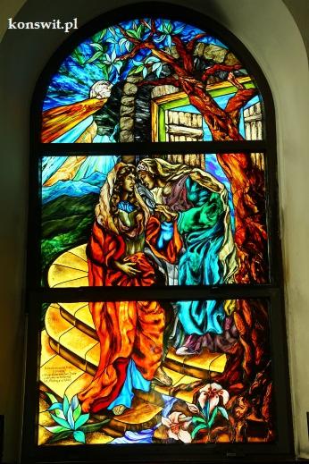 Barwny witraż przedstawiający scenę spotkania dwóch postaci kobiecych na tle pejzażu z drzewem, schodami i elementami architektonicznymi, całość utrzymana w intensywnych kolorach.