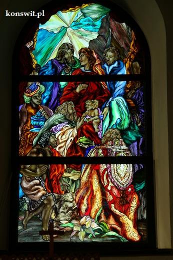 Kolorowy witraż w kościele przedstawiający scenę religijną z postaciami, w tym kobiety trzymającej dziecko, na tle abstrakcyjnych kształtów i wzorów.