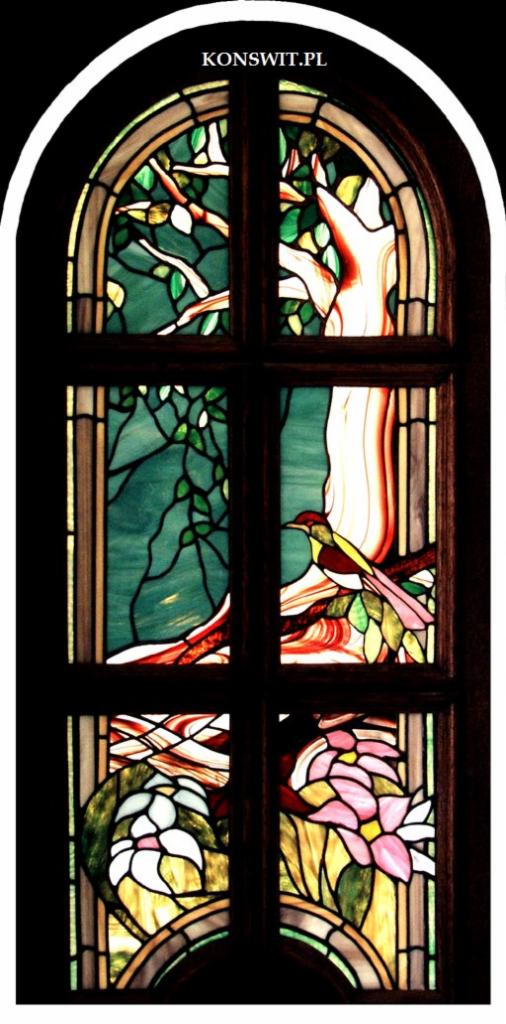 Witraż przedstawiający drzewo z ptakiem i kwiatami, w drewnianej ramie okiennej z zaokrąglonym górnym łukiem, z widocznym adresem strony internetowej w górnej części.
