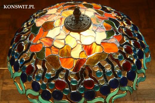 Lampa witrażowa w stylu Tiffany z dominującymi odcieniami pomarańczy, żółci, zieleni i niebieskiego, oświetlona od wewnątrz, z widocznymi detalami metalowej podstawy i wzorem przypominającym owoce.