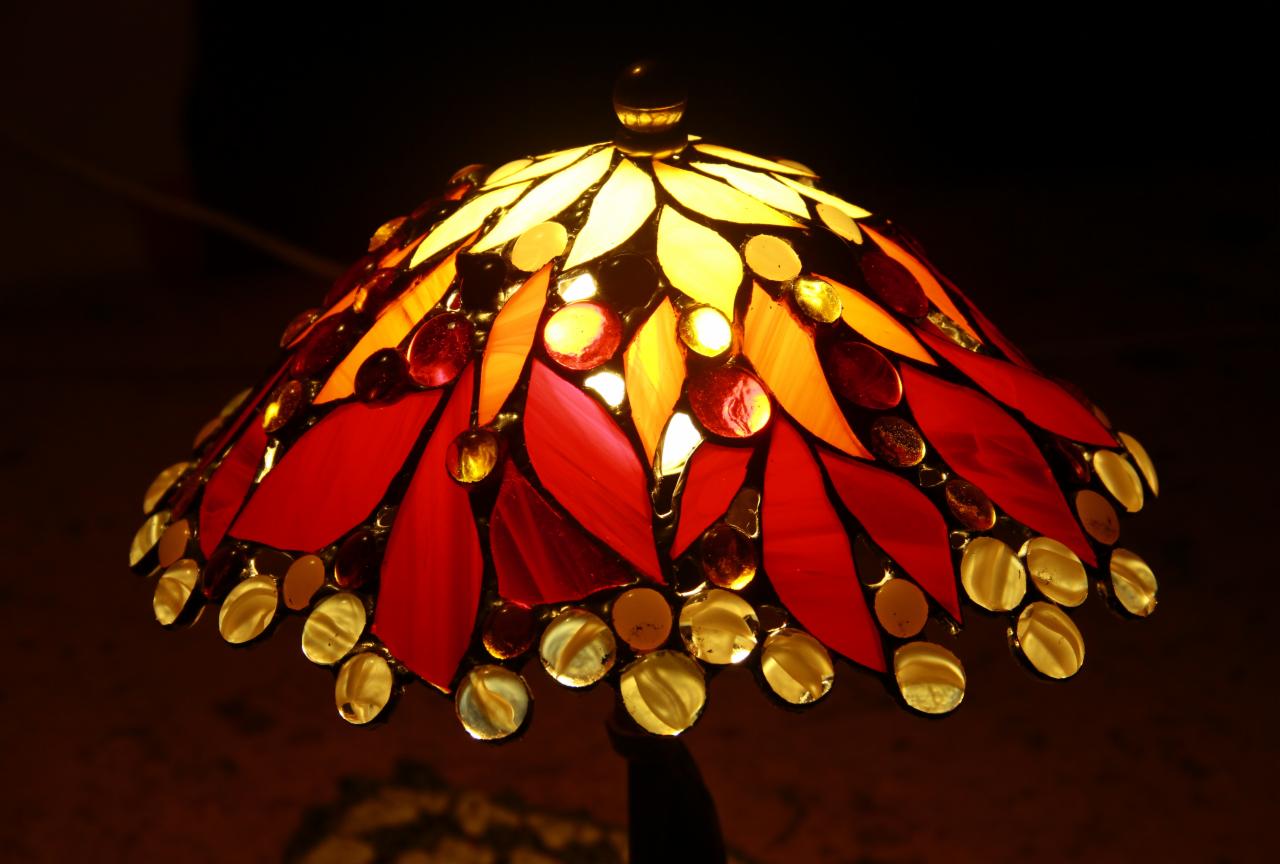 Lampa witrażowa w stylu Tiffany z czerwono-żółtymi szklanymi elementami w kształcie płatków i okrągłych paciorków, oświetlona od wewnątrz, na ciemnym tle.