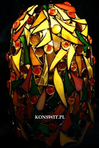 Lampa witrażowa w stylu Tiffany z geometrycznymi elementami w odcieniach żółci, czerwieni i zieleni, oświetlona od wewnątrz, z widocznym adresem strony internetowej w dolnej części.