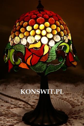 Lampa witrażowa w stylu Tiffany z motywem kwiatowym, z dominującymi odcieniami czerwieni, zieleni i żółci, umieszczona na tle ażurowej serwety, z widocznym adresem strony internetowej.