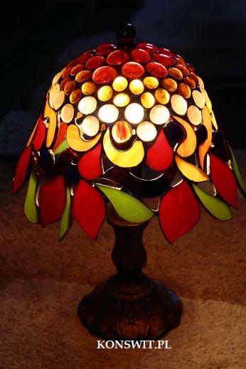 Lampa witrażowa Tiffany z motywem kwiatowym, z okrągłą podstawą i kloszem z czerwonymi, żółtymi i zielonymi elementami oraz włączonym światłem.