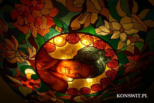 Lampa witrażowa z motywem kobiety i kwiatów w odcieniach zieleni, czerwieni i żółci, oświetlona od wewnątrz, z widocznym adresem strony internetowej w dolnym rogu.