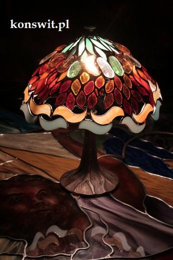 Lampa Tiffany z witrażowym kloszem w odcieniach czerwieni, zieleni i żółci, stojąca na witrażowym stole z motywem twarzy.