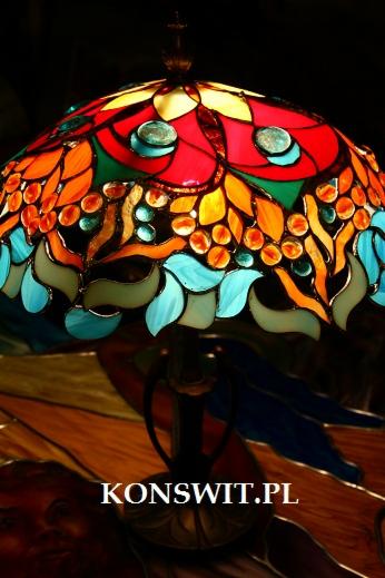 Barwna lampa witrażowa w stylu Tiffany z motywami roślinnymi, oświetlona od wewnątrz, na tle witrażowego stołu, z widocznym znakiem wodnym.