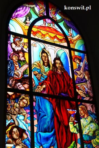 Barwny witraż przedstawiający scenę religijną z postaciami w szatach, muzykami i aniołami, umieszczony w oknie z czarnymi szprosami.