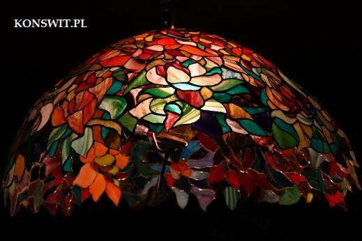 Witrażowy klosz lampy w stylu Tiffany z motywem kwiatowym, dominują odcienie czerwieni, pomarańczy i zieleni, widoczny napis KONSWIT.PL w lewym górnym rogu.