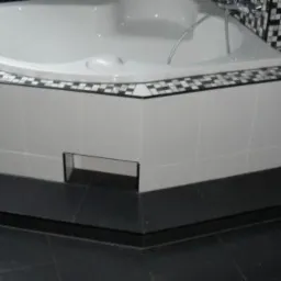 Narożna wanna z hydromasażem obudowana białymi płytkami z czarno-białym mozaikowym wykończeniem, posadzka wyłożona czarnymi płytkami, czarna ściana z wzorem.