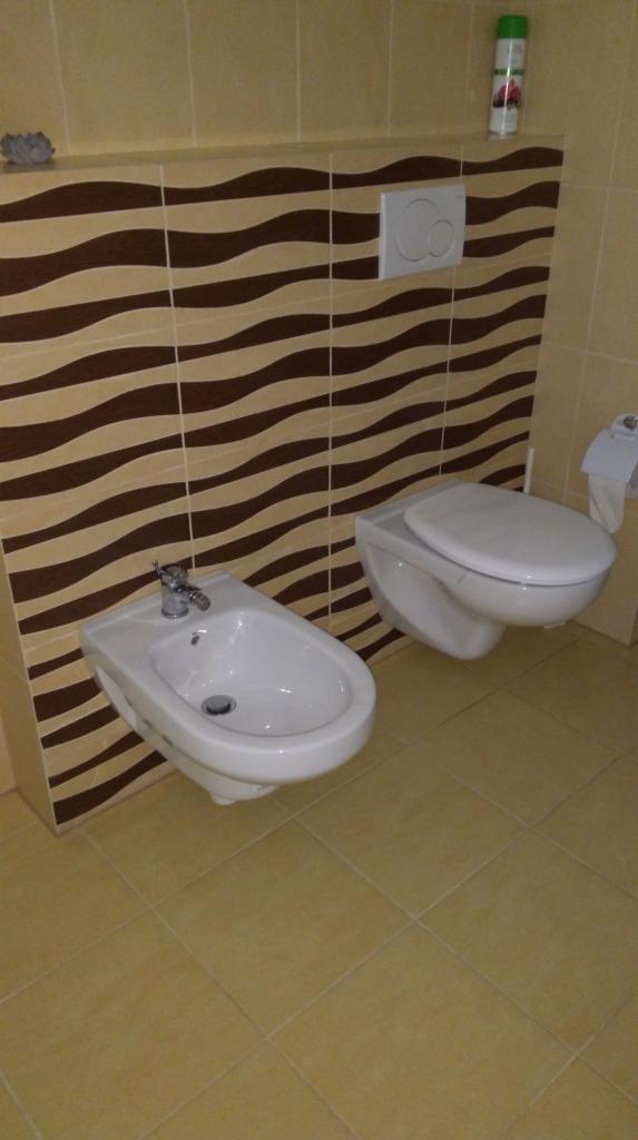 Bidet i podwieszana toaleta w łazience z brązowo-beżowymi płytkami w falisty wzór na ścianie, kremowe płytki podłogowe, widoczny dozownik mydła i odświeżacz powietrza.
