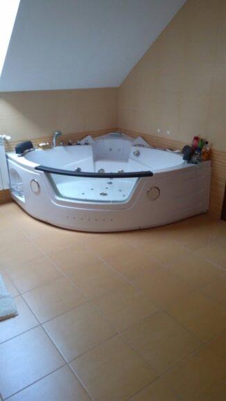 Narożna wanna z hydromasażem w łazience na poddaszu, beżowe płytki podłogowe i ściany, w tle widoczny grzejnik.