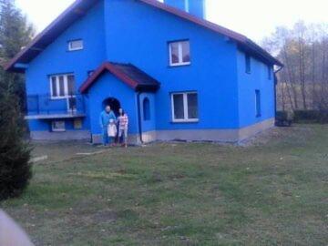 Dom jednorodzinny po remoncie elewacji, pomalowany intensywnym, jednolitym błękitem, z widoczną rodziną stojącą przed wejściem. Czerwone obramowanie wejścia i dachu kontrastuje z błękitną elewacją.