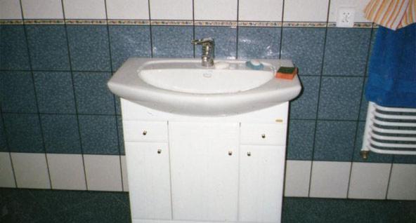 Biała umywalka z szafką w łazience z niebieskimi płytkami, widoczny fragment grzejnika i kontakt.