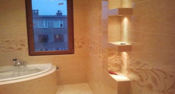 Beżowa łazienka z wanną narożną, oknem z widokiem na blok i wnękami z oświetleniem punktowym, dekorowana motywem roślinnym na płytkach.