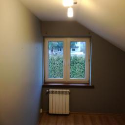 Private - Pokój na poddaszu po remoncie: szare ściany, drewniana podłoga, białe okno z widokiem na zieleń, biały grzejnik pod oknem, kinkiet na ścianie.