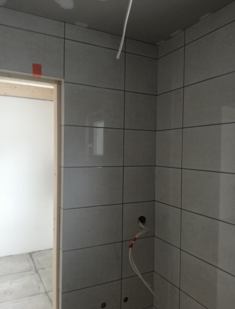 Świeżo ułożone, błyszczące, szare płytki ceramiczne na ścianach łazienki w trakcie remontu, widoczne otwory na instalacje i wystające przewody, zabezpieczone taśmą. Fragment futryny drzwi bez...