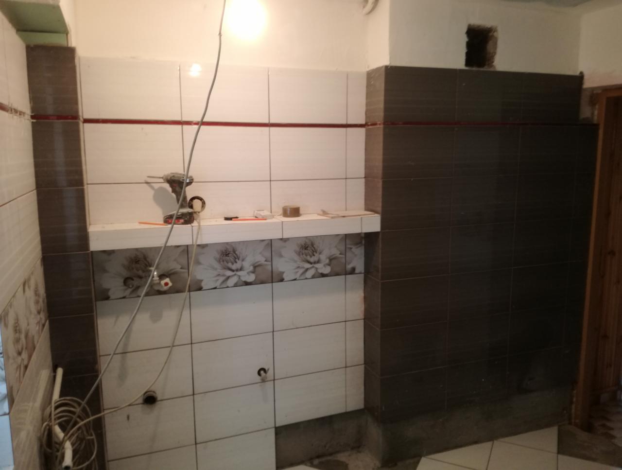 Układanie płytek w łazience: białe i brązowe płytki, narzędzia glazurnicze na półce, widoczne przewody elektryczne i niedokończone fugowanie.