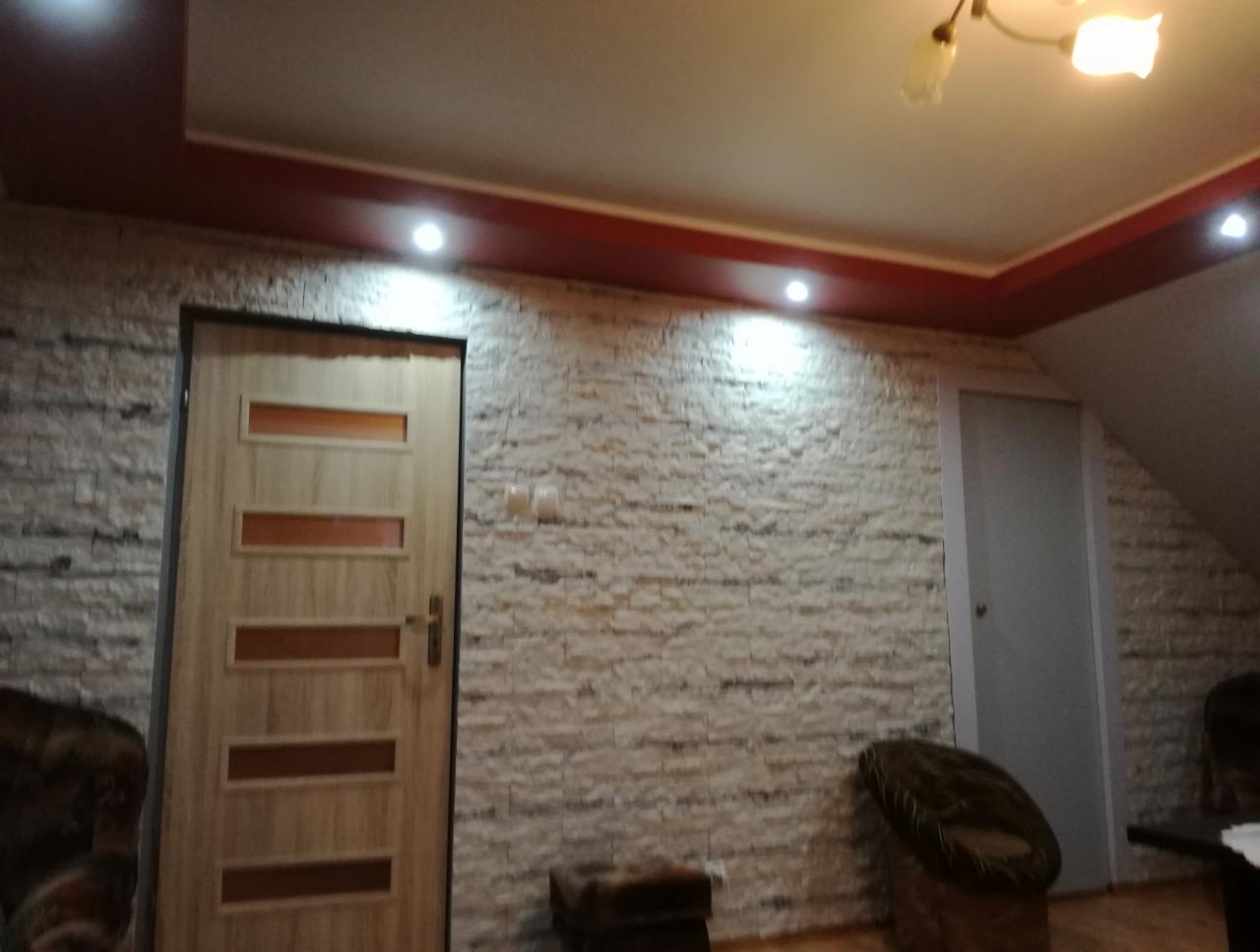 Ściana wyłożona dekoracyjną cegłą w odcieniach bieli i szarości, z drewnianymi drzwiami z przeszkleniami i białymi drzwiami na poddasze, oświetlona punktowymi lampami i lampą wiszącą.