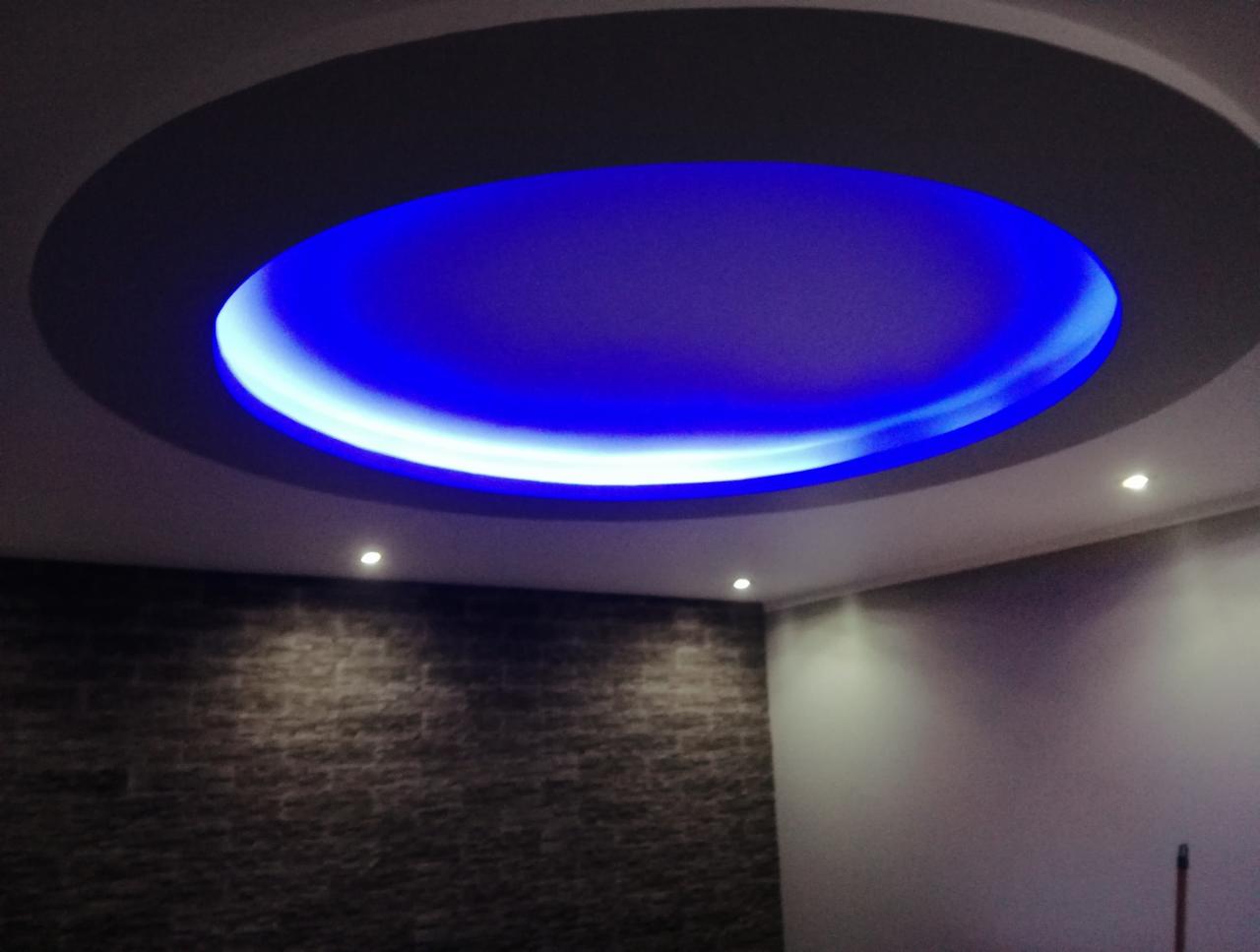 Okrągły podwieszany sufit z niebieskim oświetleniem LED, widok z dołu, ściana z dekoracyjną okładziną kamienną w tle.